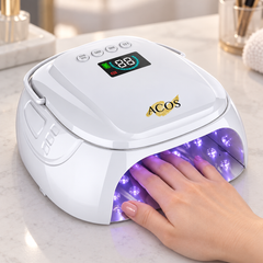 A white ACOS UV/LED nail lamp with a digital display and control buttons, shown with a hand resting inside the illuminated purple lamp on a marble countertop.